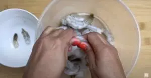 Hands peeling shrimp over a bowl on a wooden countertop.